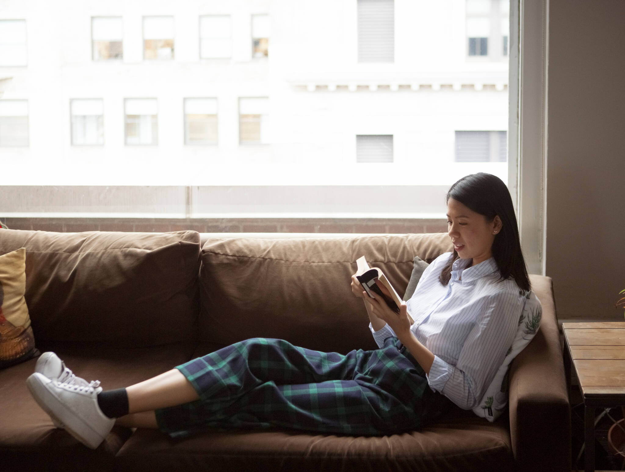 Side View Of Businesswoman Reading Book While Relaxing On Sofa By Window In Office