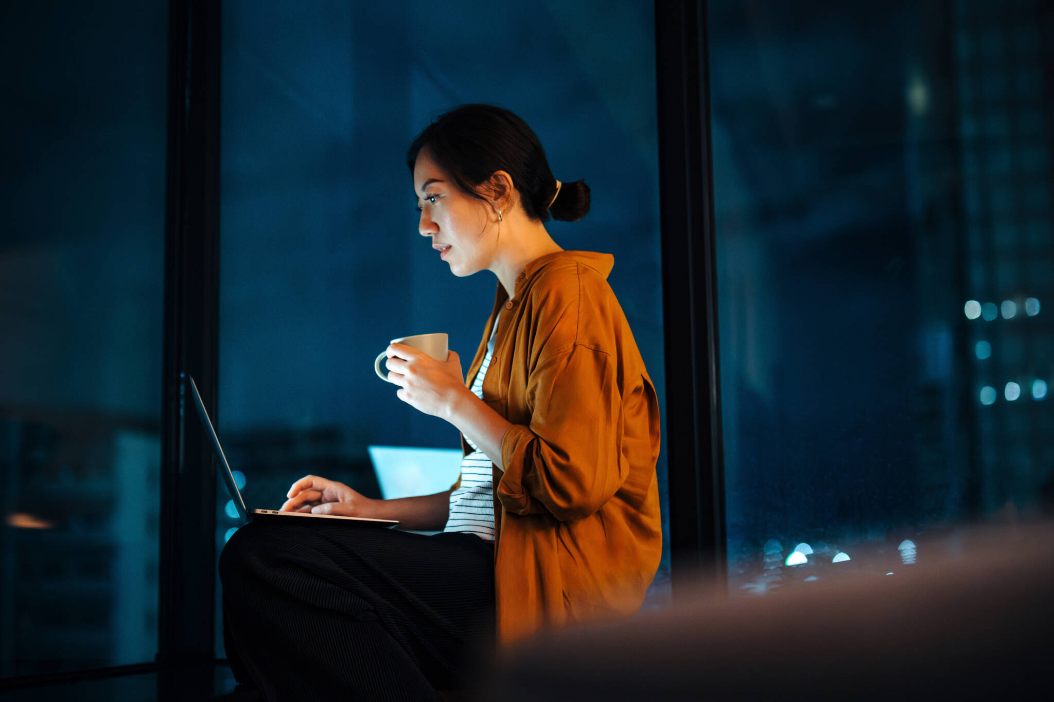 Young Asian Woman Sitting By The Window Against Illuminated City Scene In The Dark, Working On Laptop In The Living Room At Home. Working From Home. Freelancer. Self Employment. Business Lifestyle And Technology